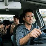 Driver focused ahead while passengers talk in the background.